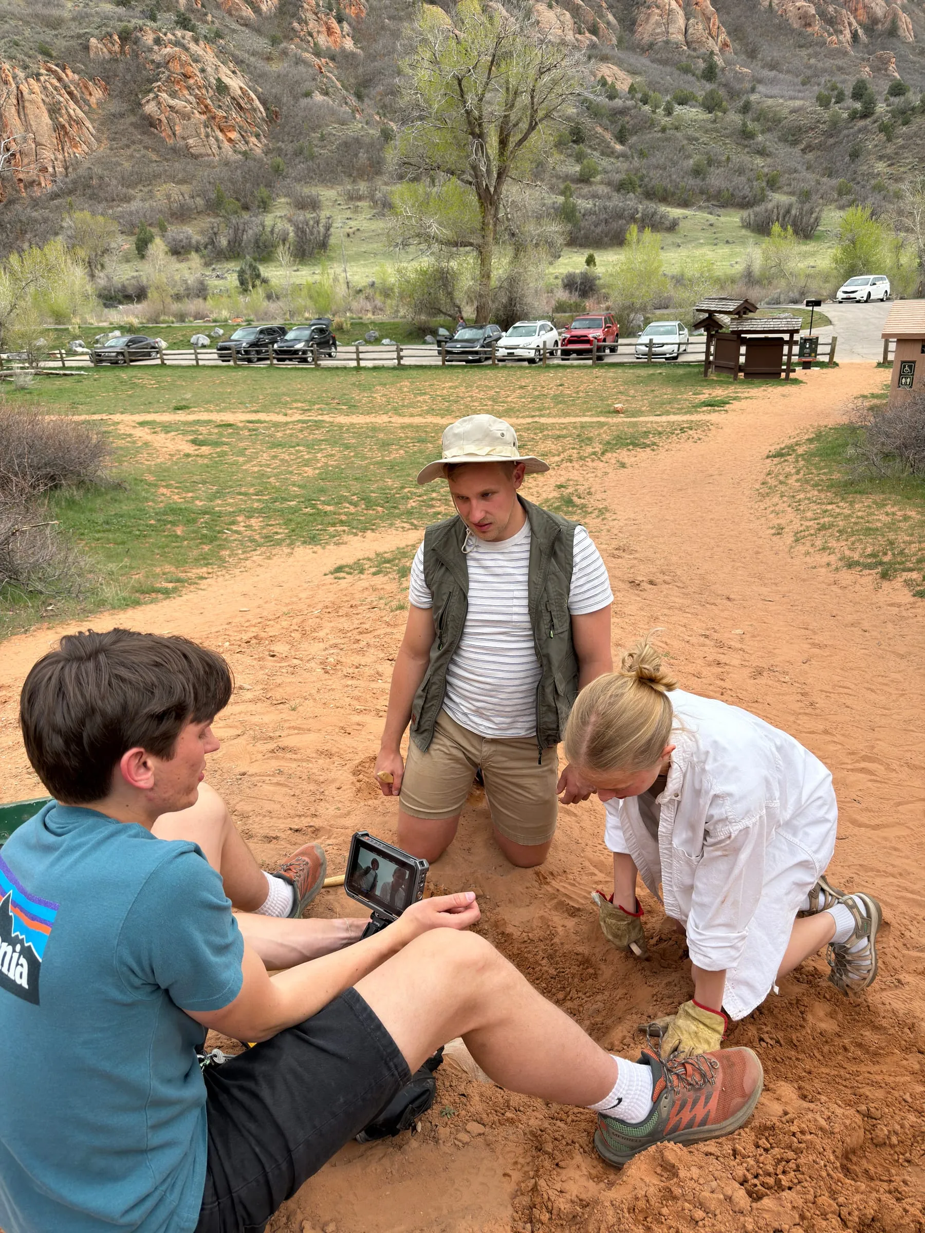 On a red sand trail, two actors kneel at the dig while a crew member watches a small monitor in front of them