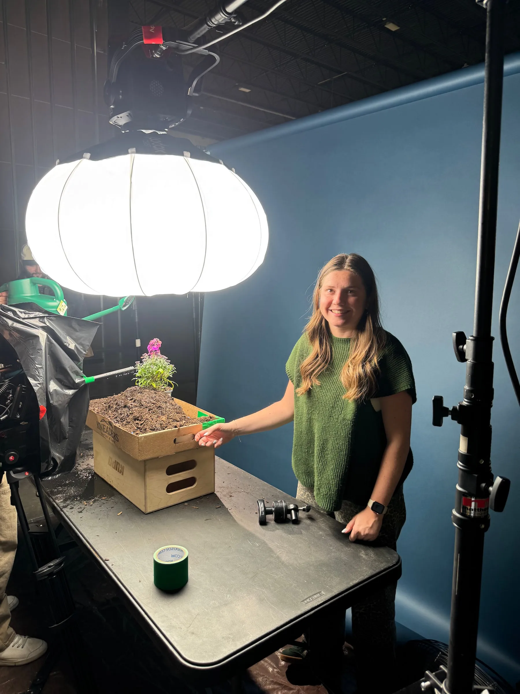 On a studio tabletop set, a woman smiles beside a flowering plant in a box of soil under a big lantern light