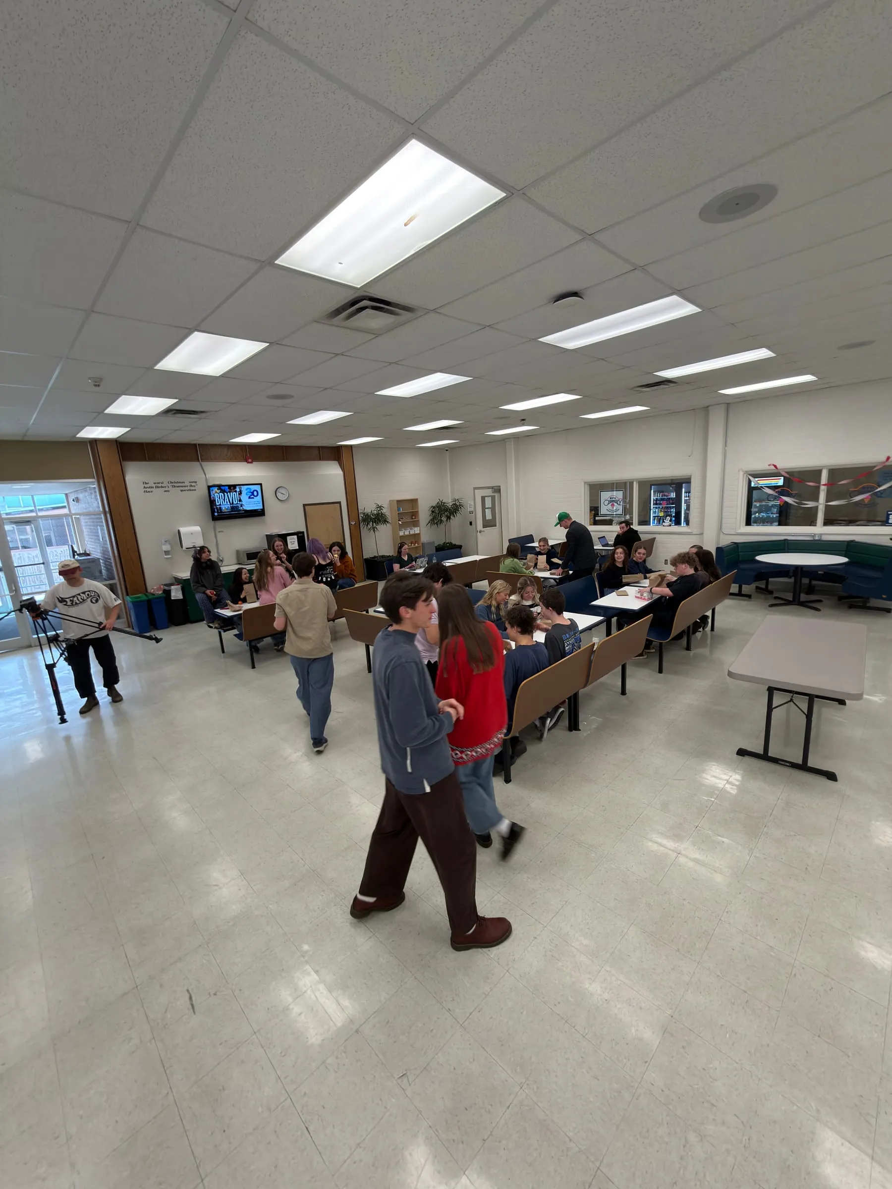 In a school lunchroom, a camera operator sets up on a slider as students walk between the tables