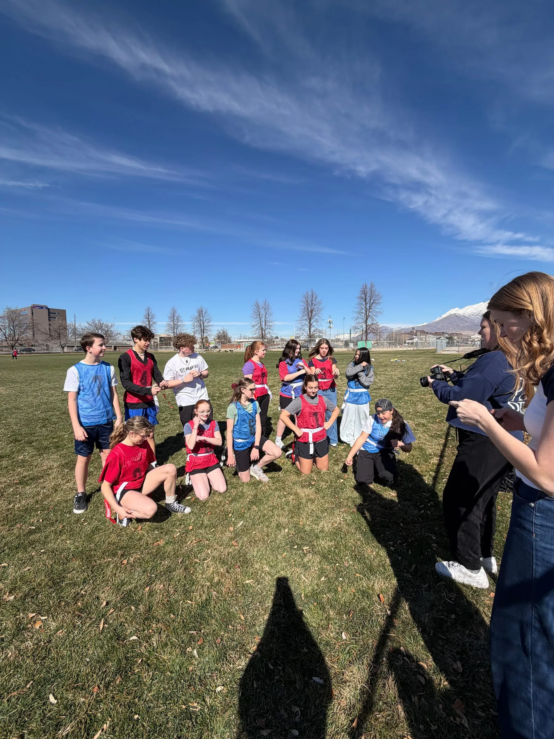 Teens in red and blue flag football pinnies gather on a field while a camera operator films from the side