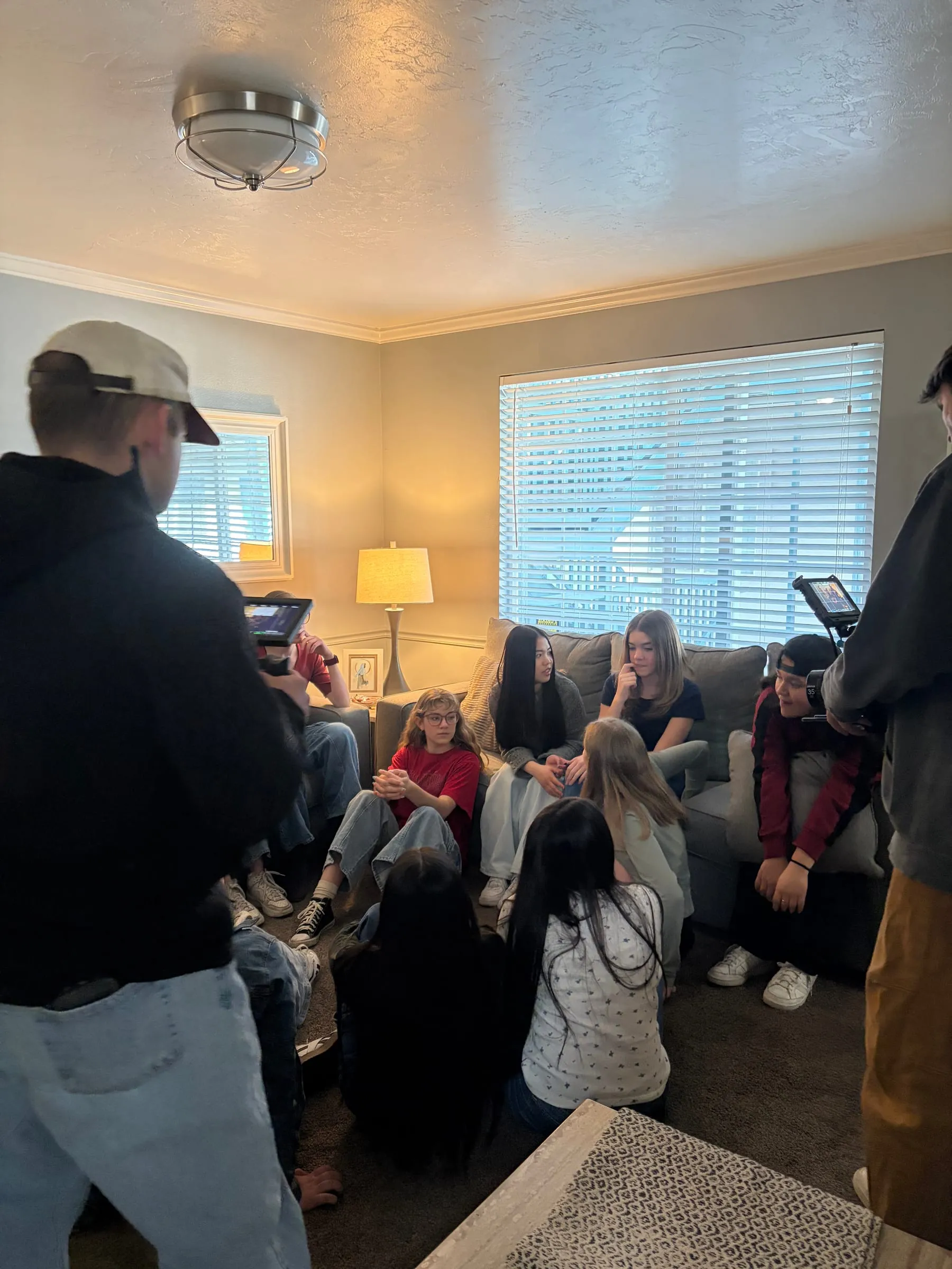 Crew with monitors in the foreground film a group of teens packed onto the couches and floor of a living room