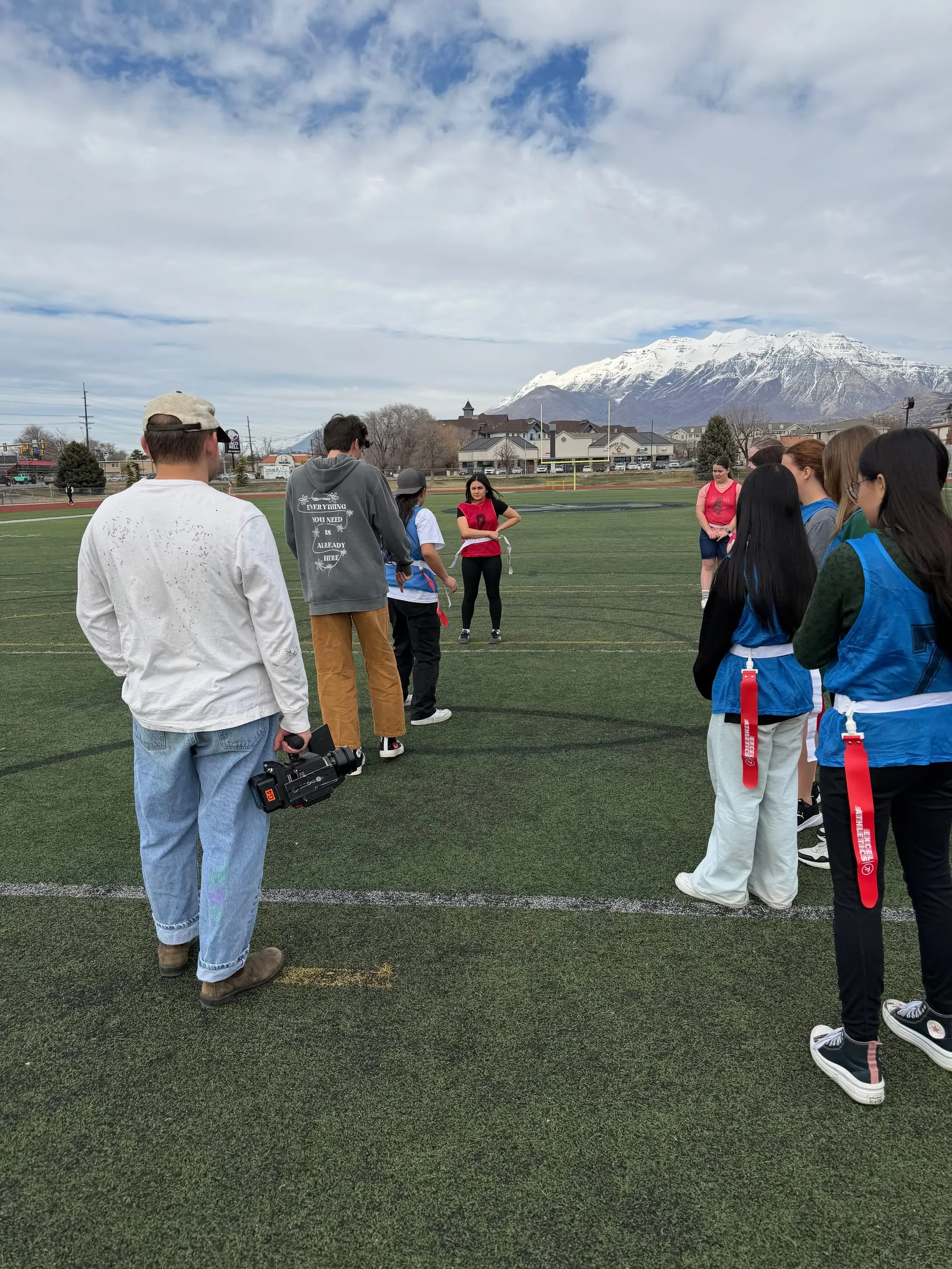 On a turf field under a snowy mountain, the crew sets up a shot with teens in pinnies and flags