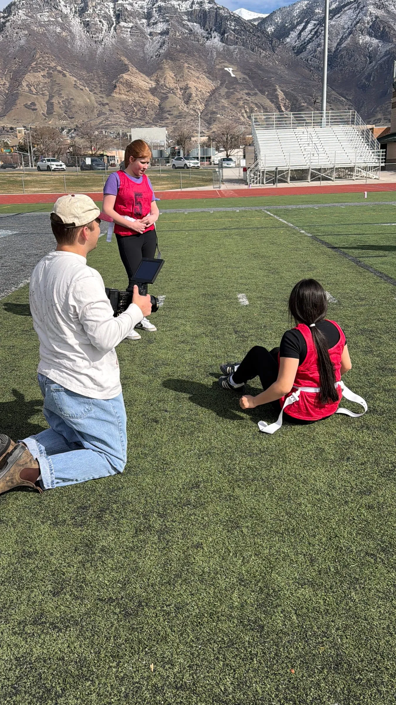 Kneeling on the turf, a crew member films a girl in a red pinnie sitting on the field