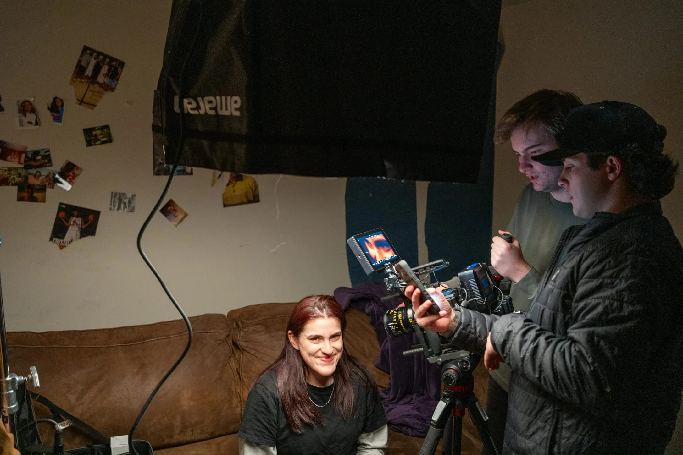 In the basement set, with photos taped to the wall, two crew members check a camera on a tripod while an actor smiles from the couch