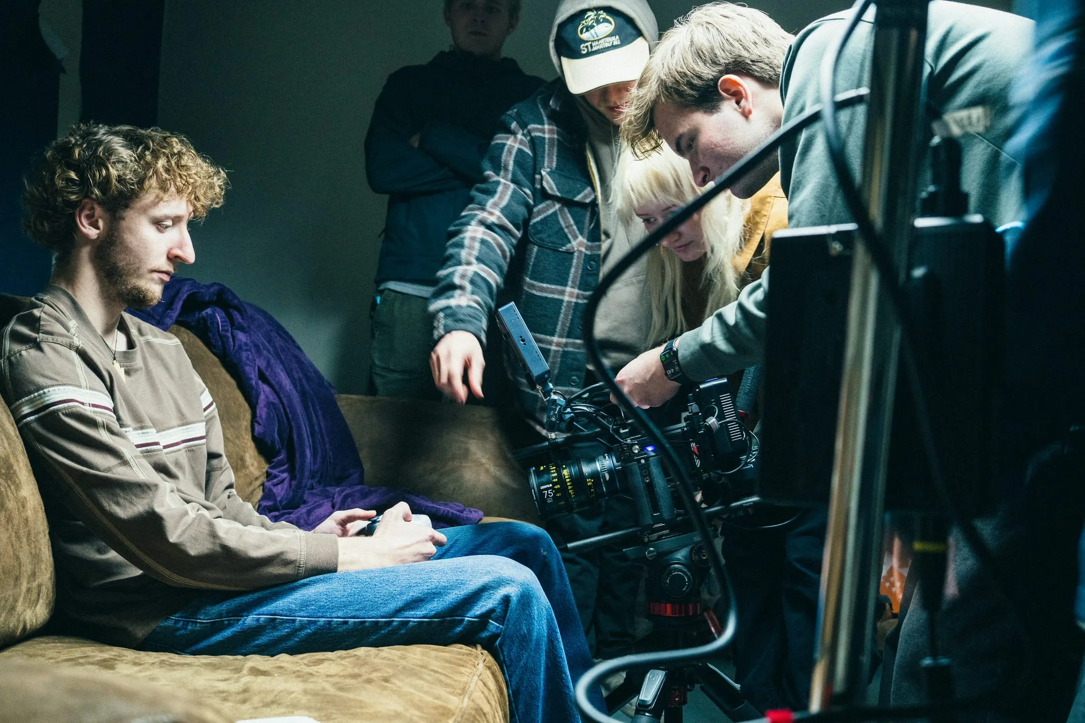 An actor holds a controller on the couch while the crew huddles over the camera beside him