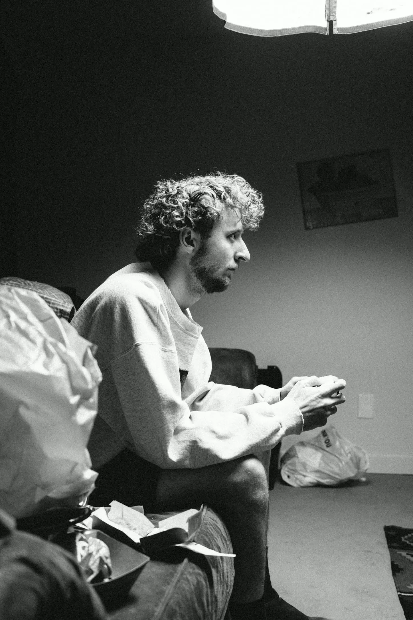 In black and white, an actor sits playing a video game under a single light, trash bags at his feet