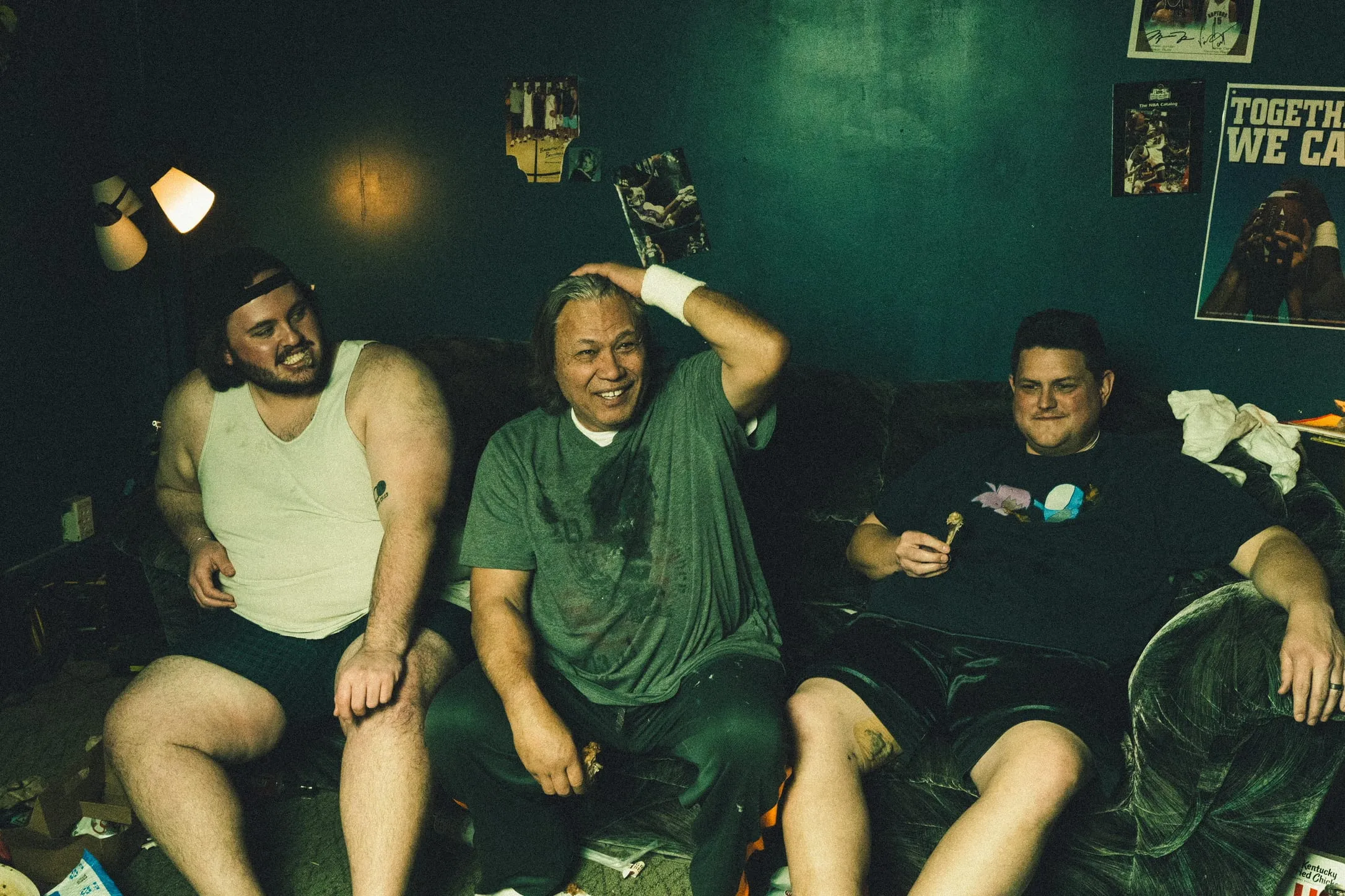 Three men laugh on a couch in a green room hung with sports posters