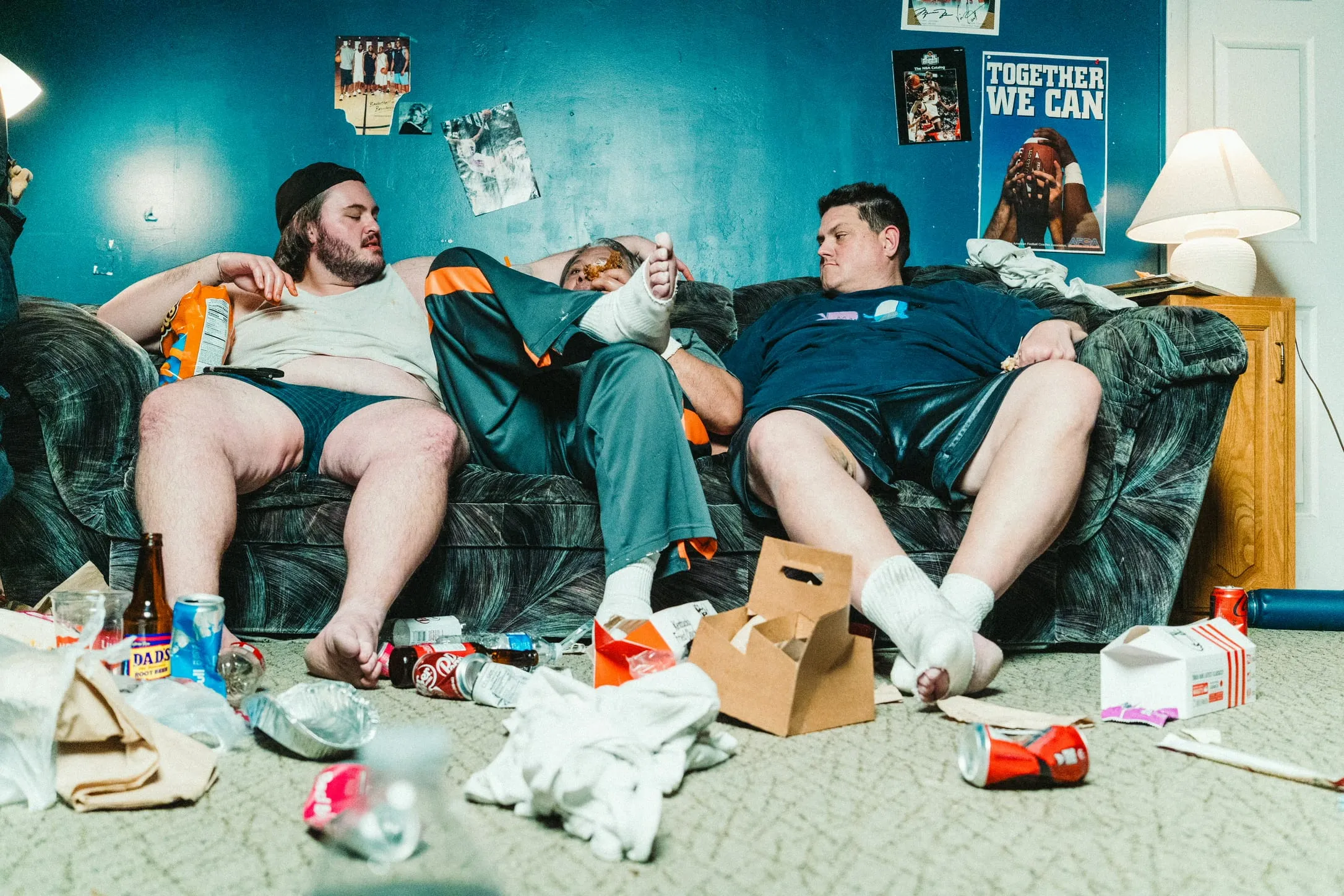 The same three men sprawl on the couch in a room littered with cans, wrappers and takeout