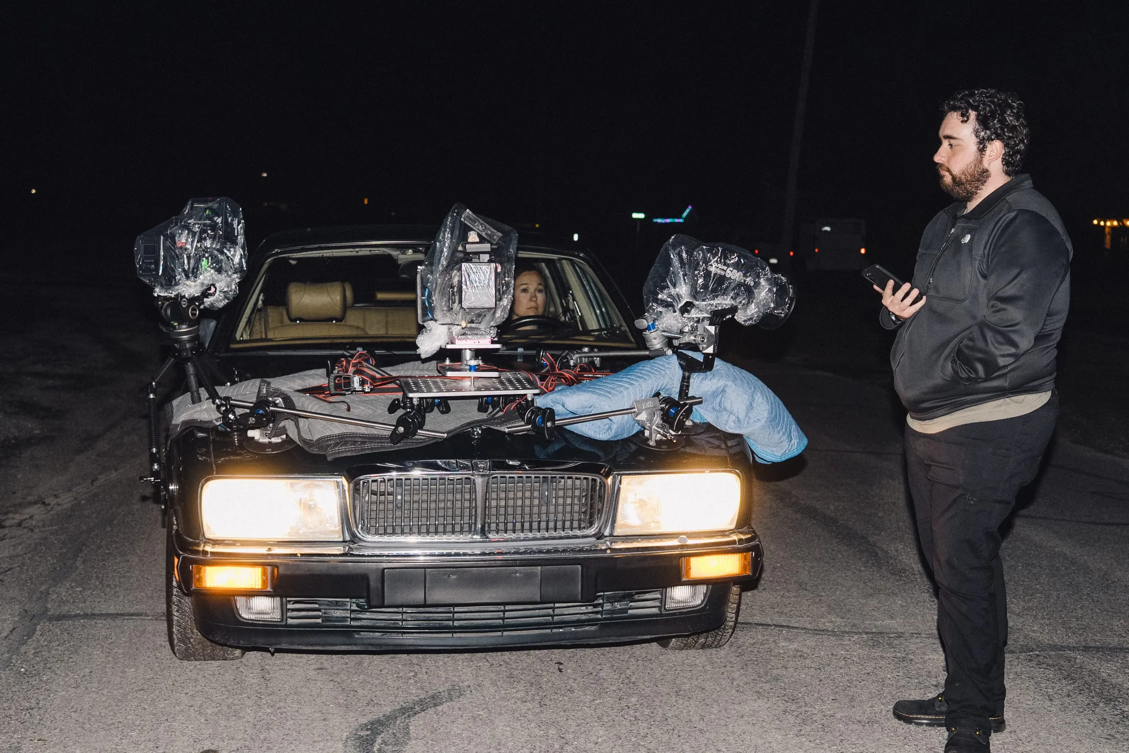 At night, cameras in rain covers sit rigged on the hood of a car with its headlights on while a crew member stands beside it