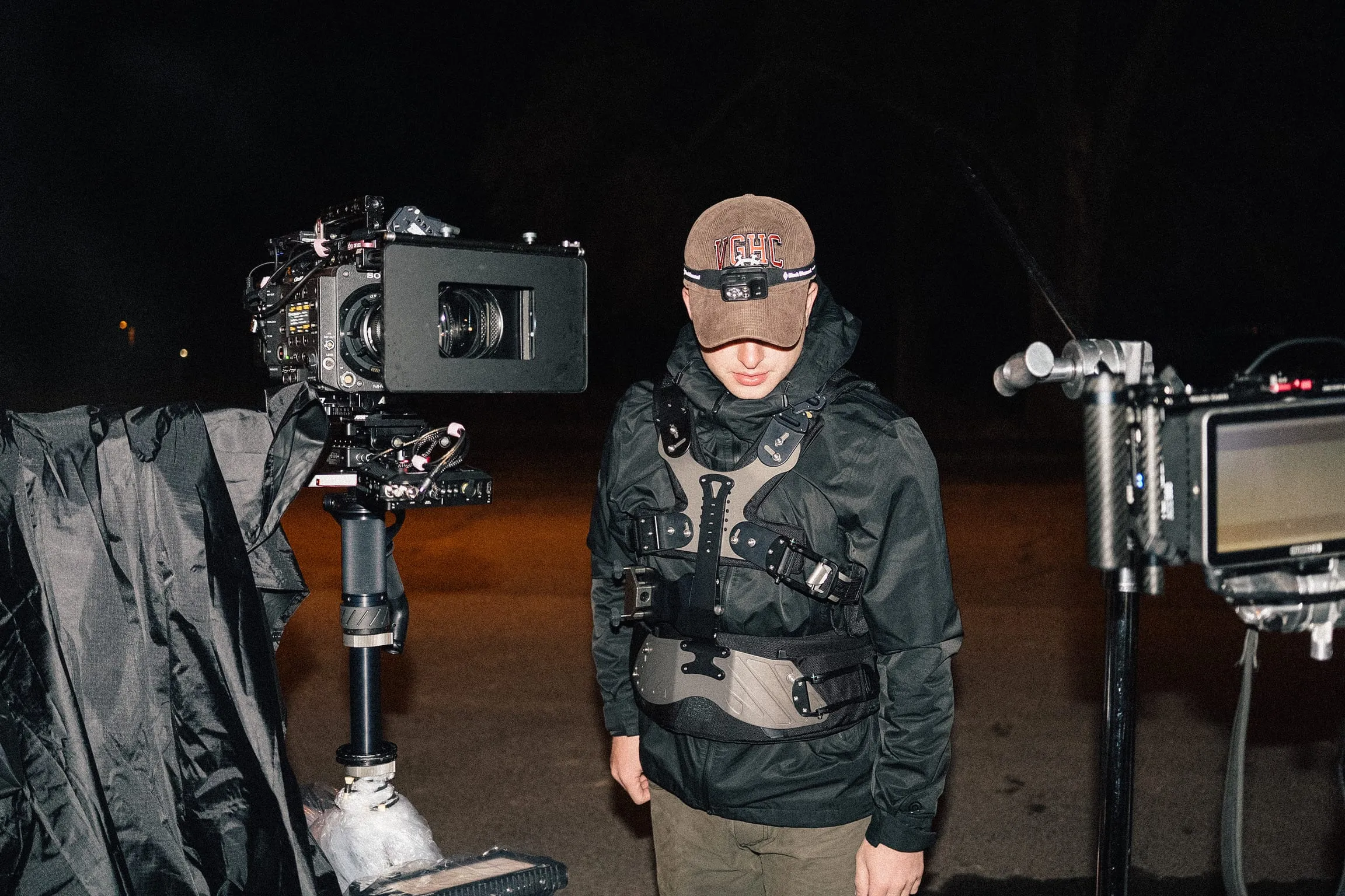 At night, a camera operator in a stabilizer vest stands between the camera arm and a monitor