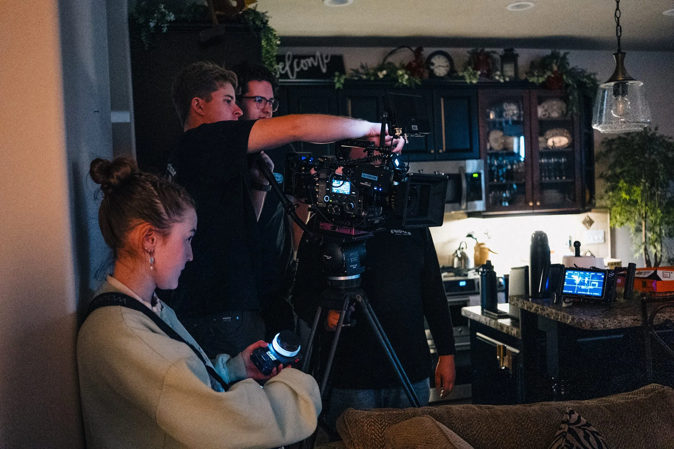 In a kitchen, a camera assistant adjusts focus while the operator frames the shot