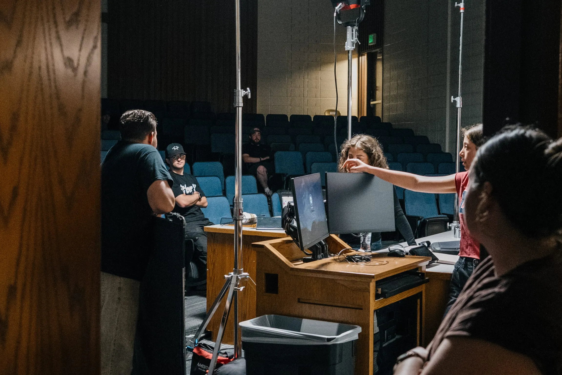 In a lecture hall, the crew works from a podium with monitors as one of them points out the setup