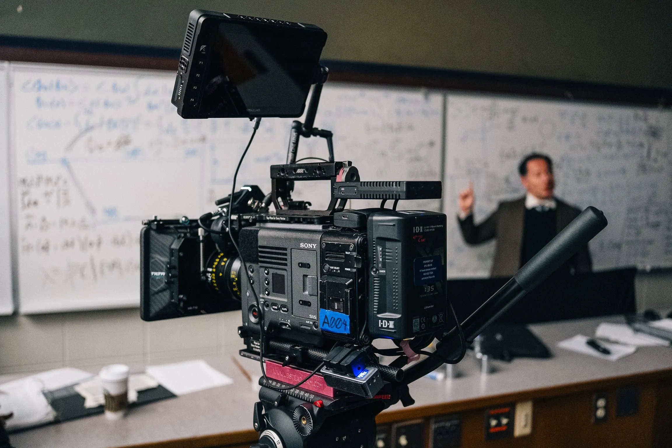 A camera and monitor in the foreground while an actor in a suit lectures at a whiteboard full of equations