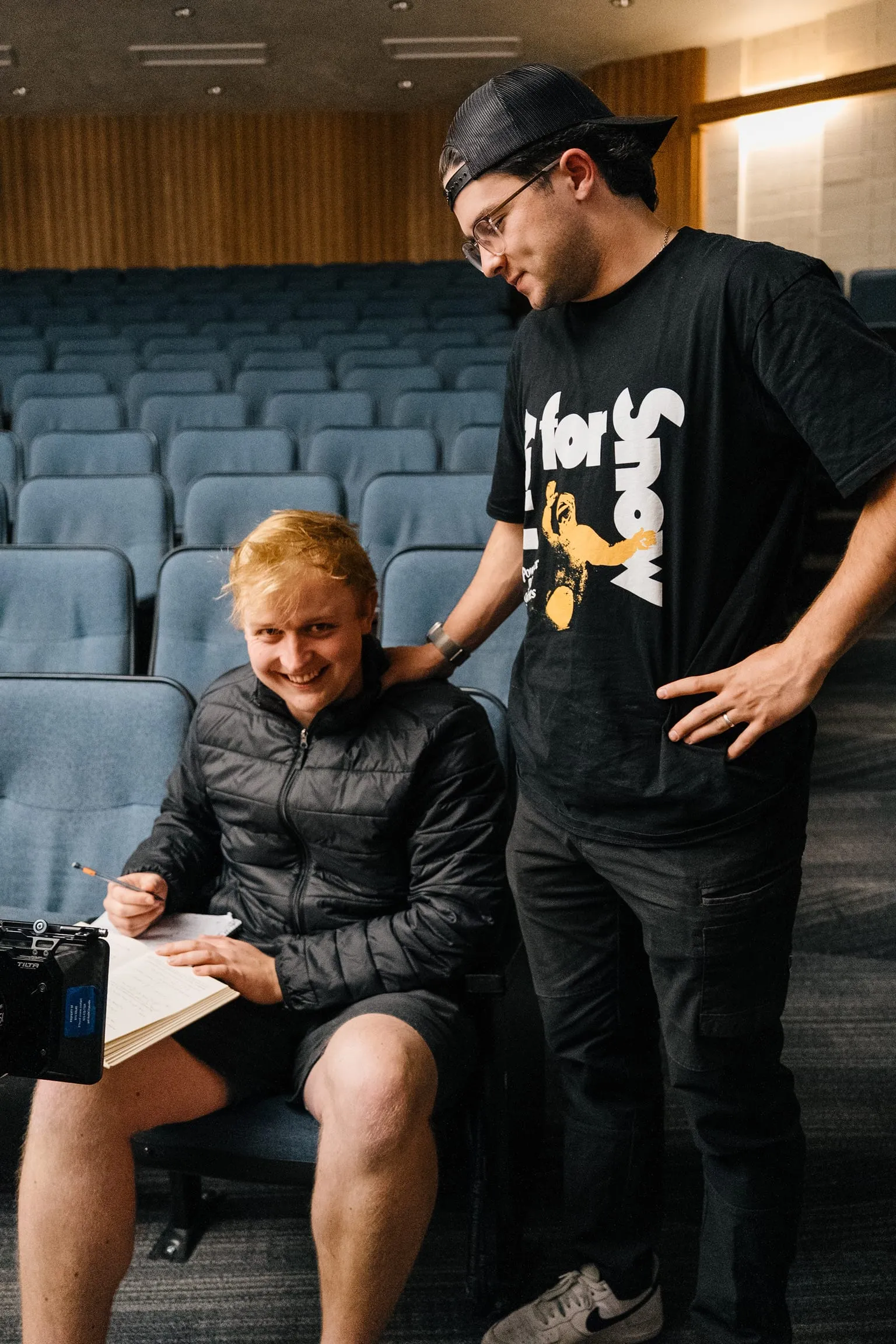 In the lecture hall seats, a crew member with a script grins while another leans over his shoulder