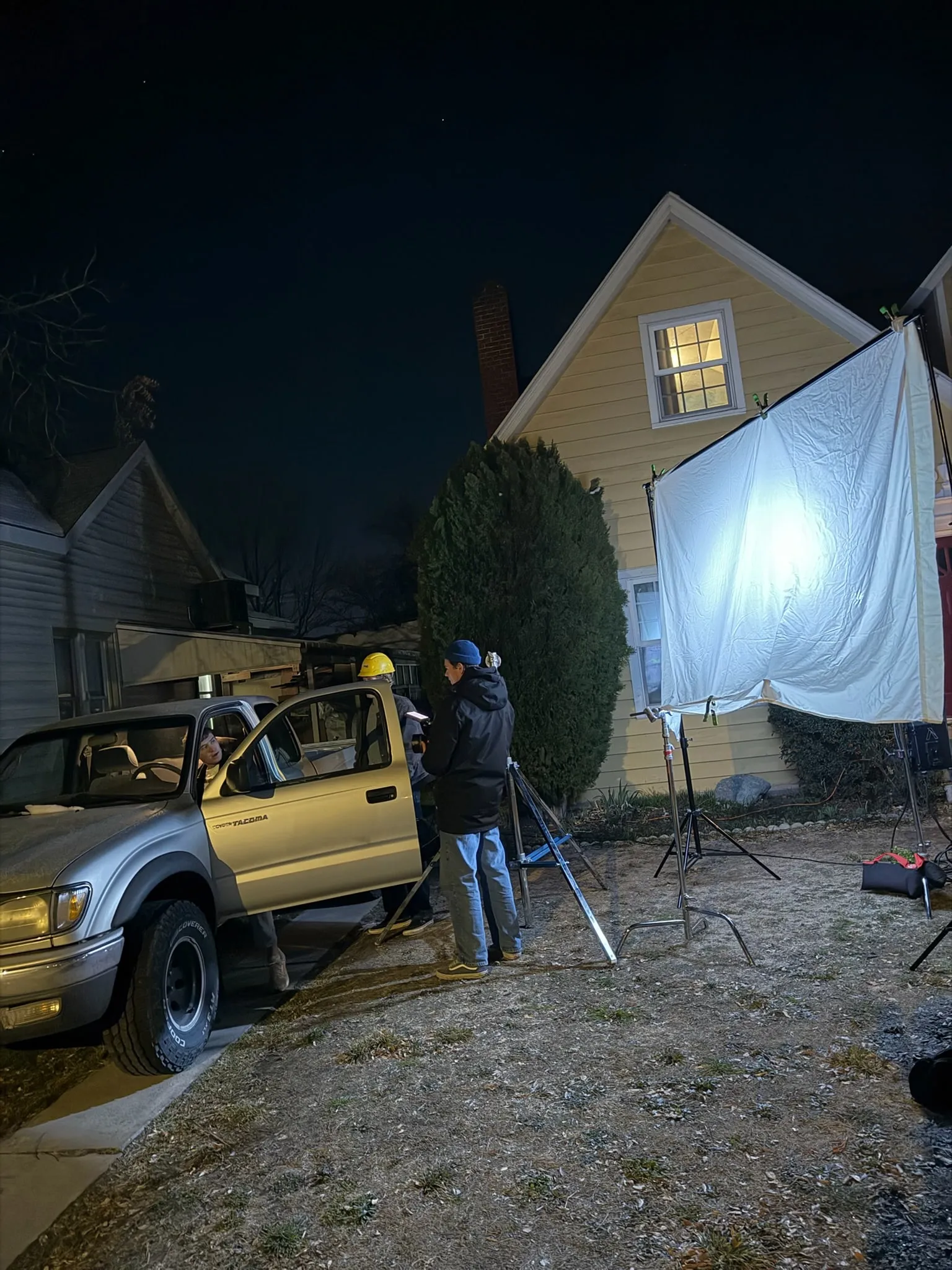 At night outside a house, a big diffusion frame glows as a camera operator films a young man sitting in a pickup with its door open