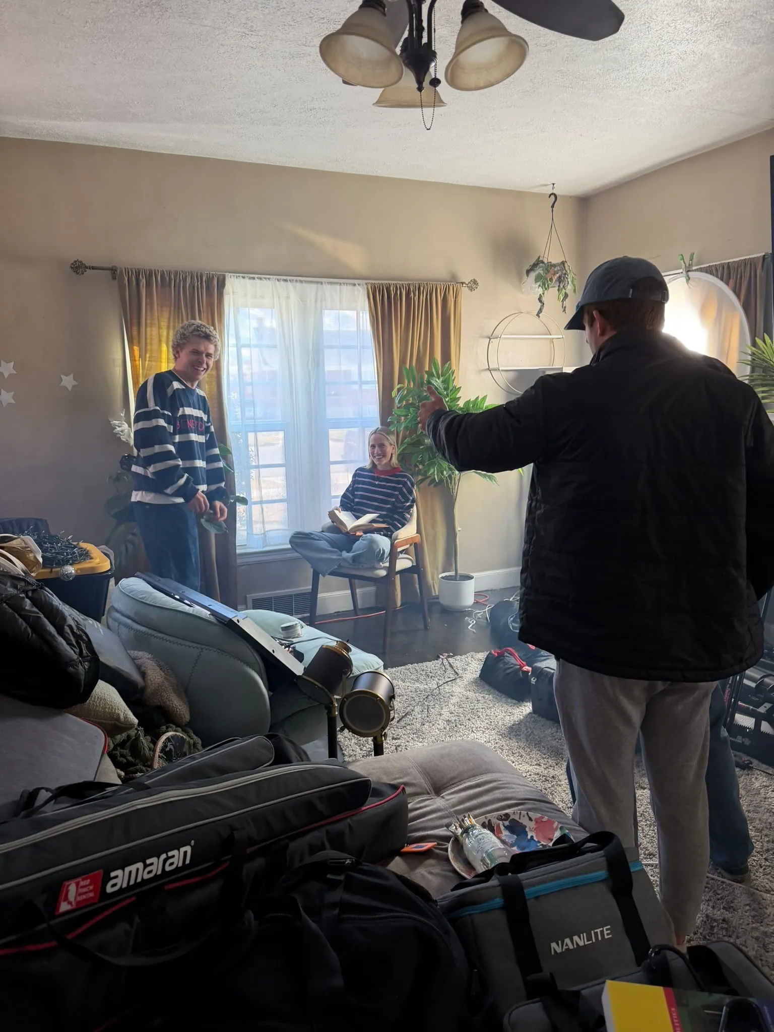 In a sunny living room, a man in a cap gestures to a laughing young man and a young woman reading in a chair, gear bags piled in front