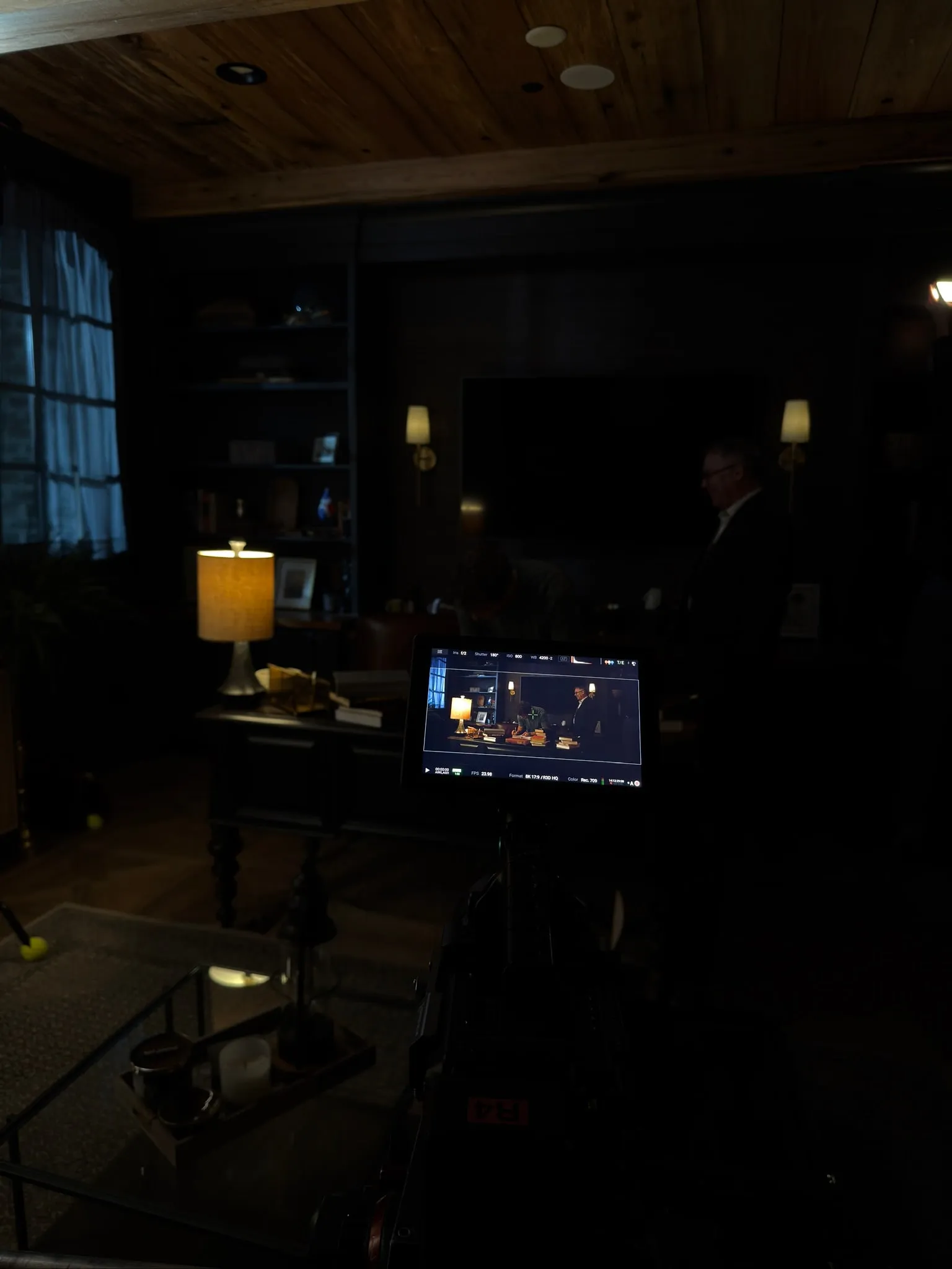 In a dark study, the camera monitor shows the scene: a young man writing at a desk of books and an older man standing behind him