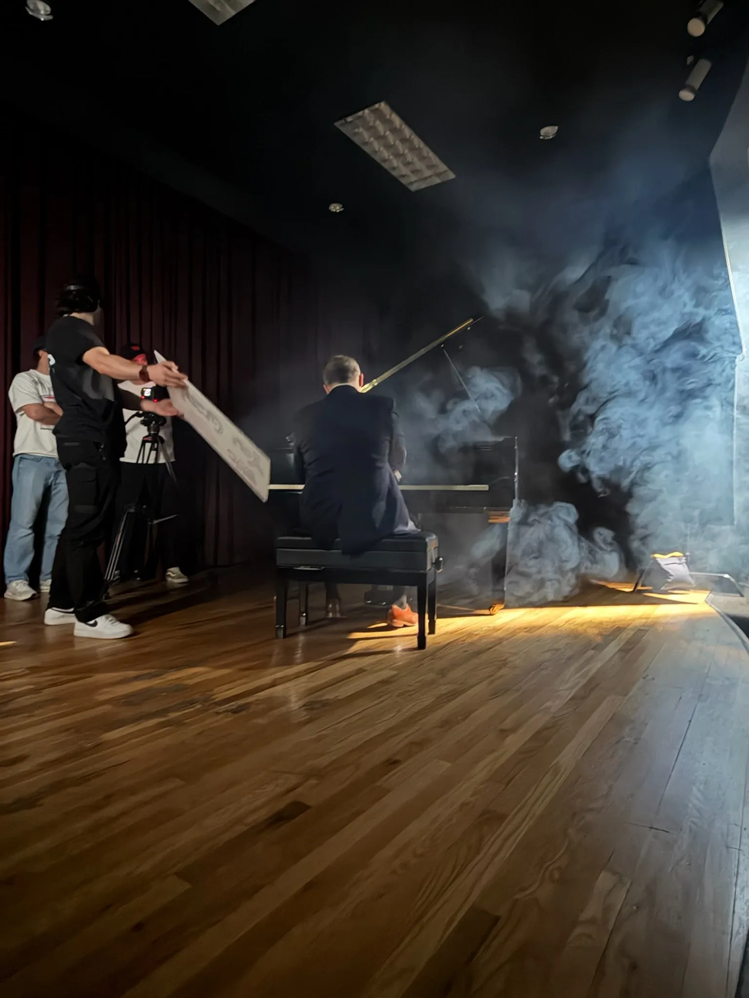 On a hazy stage, a man in a suit sits at a grand piano, seen from behind, as crew hold a bounce board and film from the side