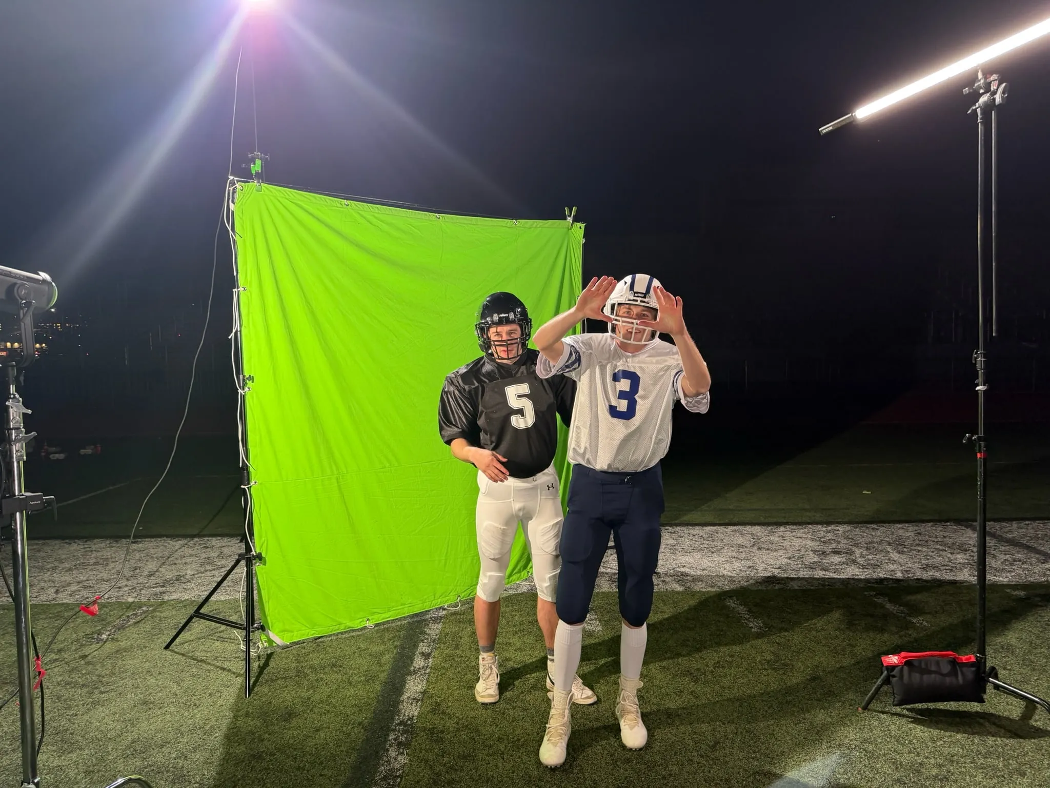 At night on a turf field, two football players, number 3 and Tommy in number 5, pose in front of a green screen between big lights