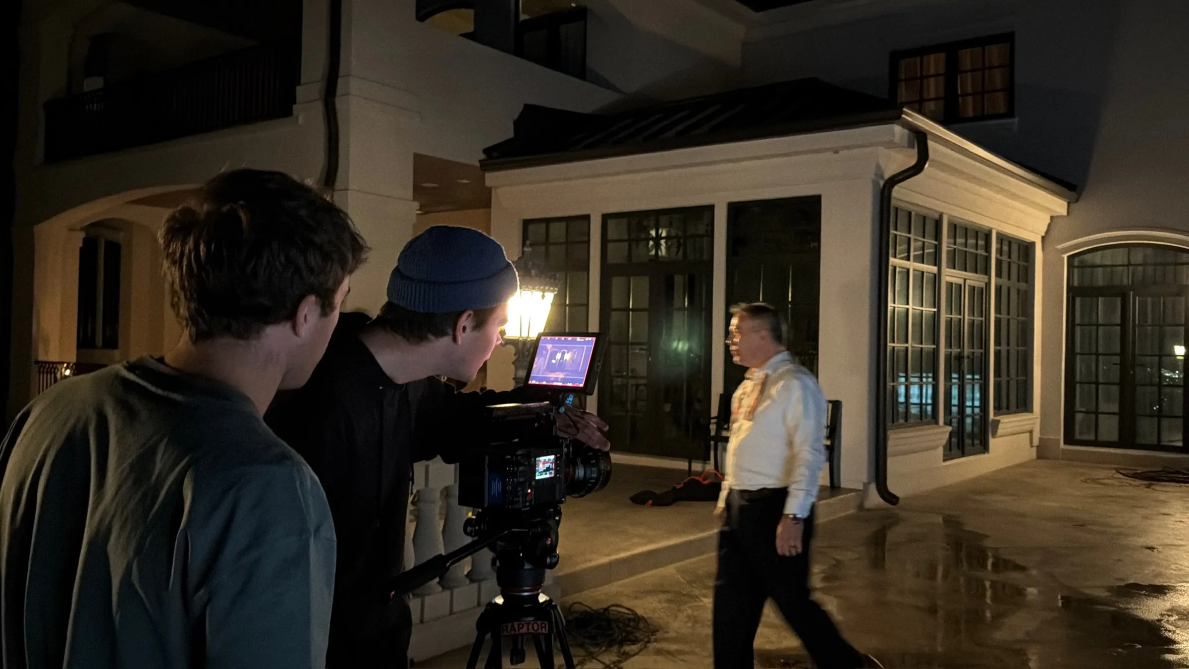 At night on a wet patio, a camera operator watches the monitor as an older man in a white shirt walks through the shot