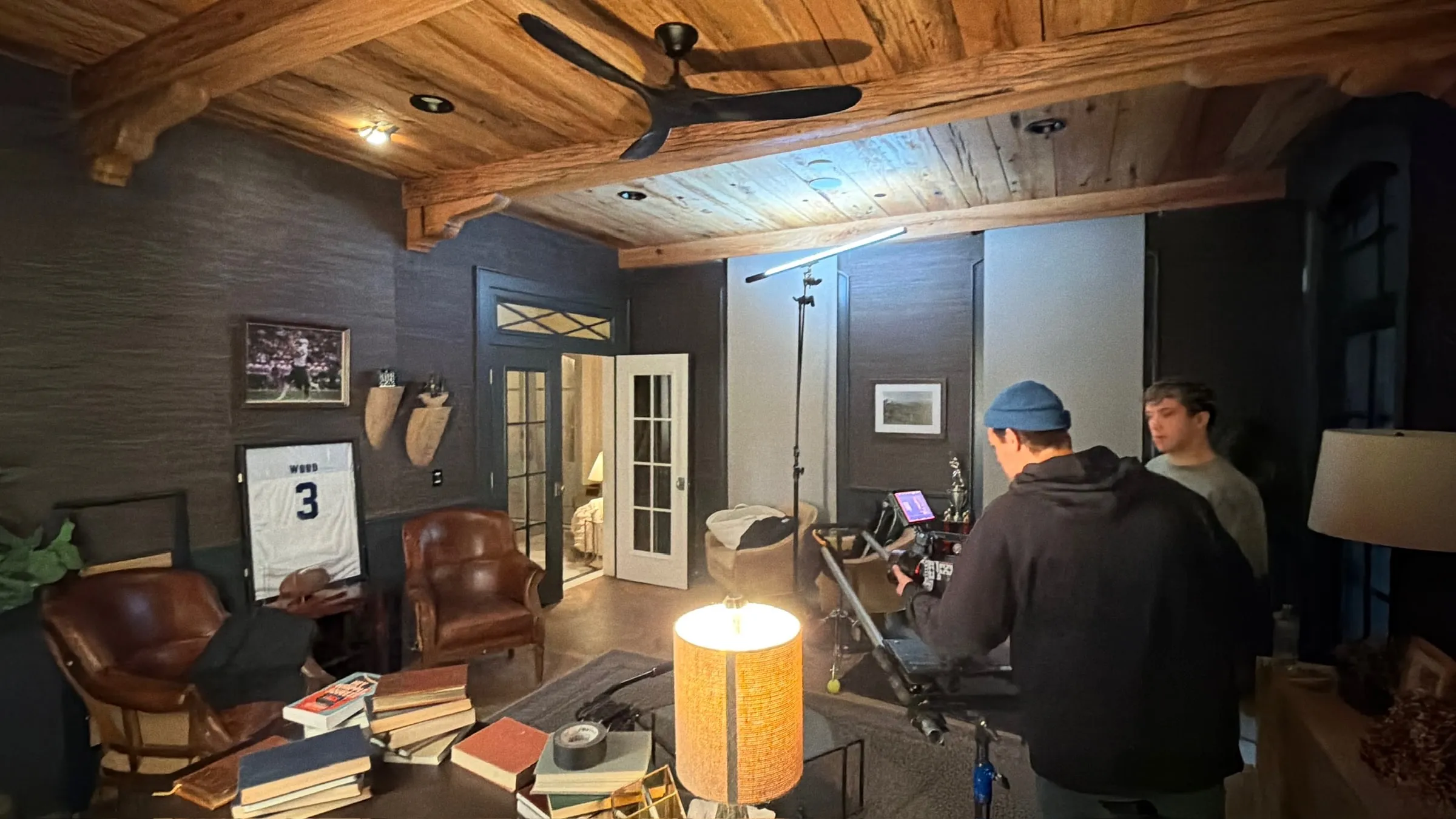 A camera operator lines up a shot in the study set, a framed number 3 jersey on the wall behind leather chairs