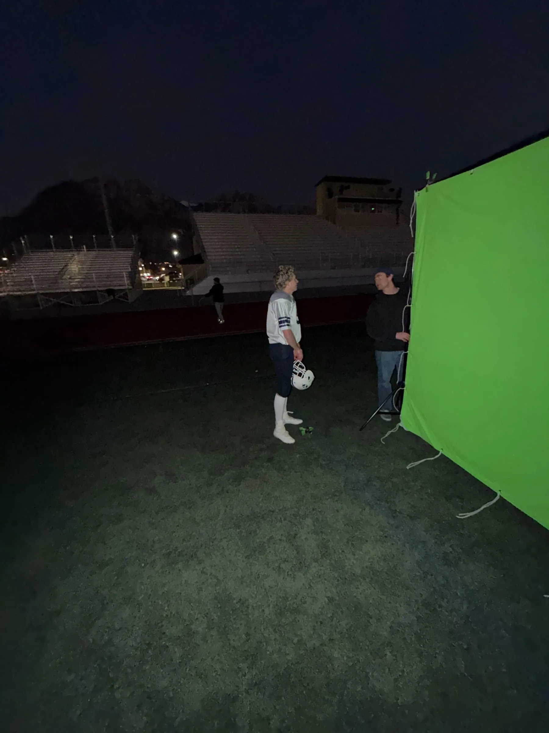At night on a football field, a player holding his helmet talks with a crew member beside a green screen