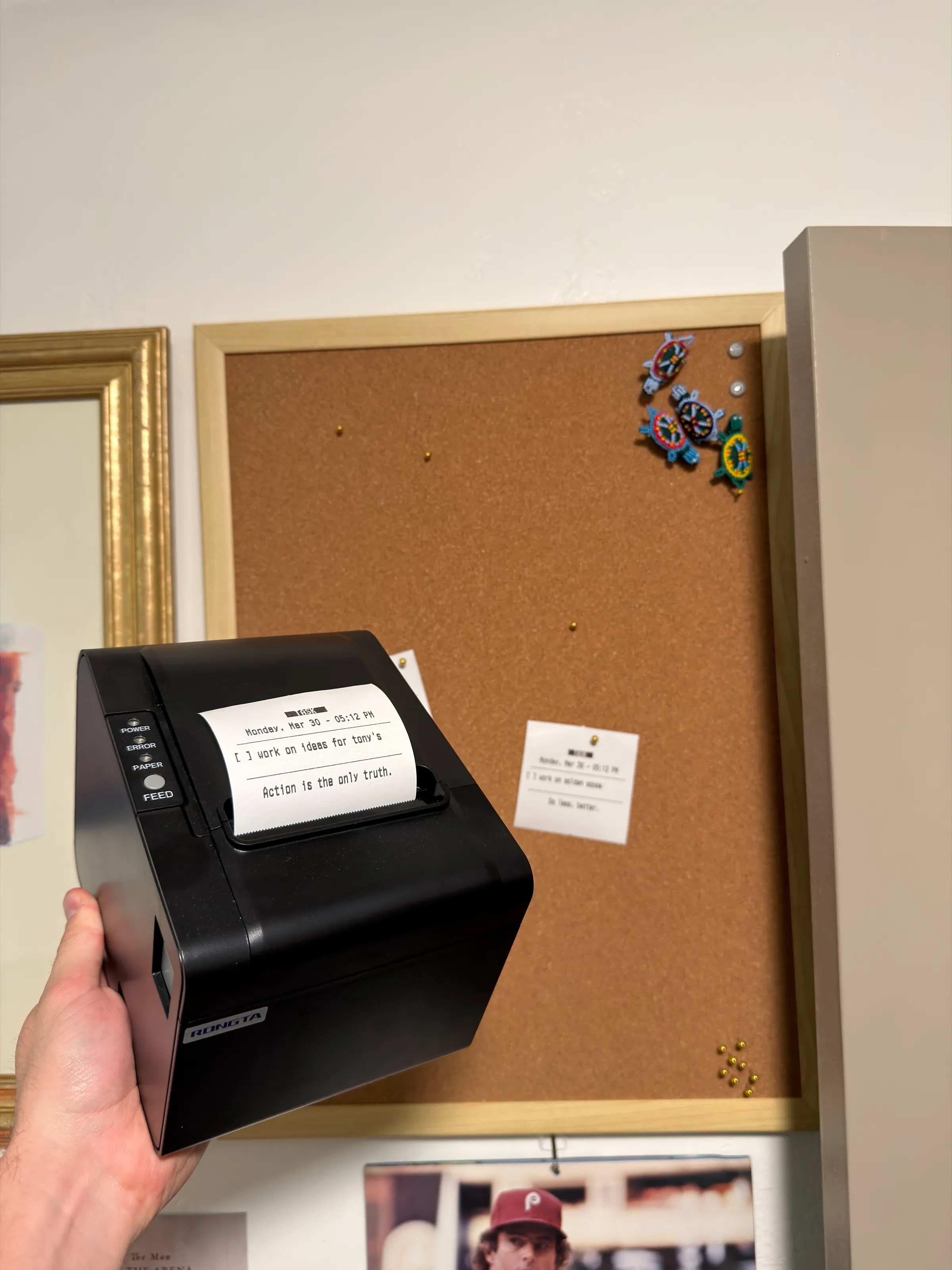 The receipt printer held up in front of a corkboard. Its receipt reads “TASK”, “Monday, Mar 30 - 05:12 PM”, a checkbox for “work on ideas for tony's” and “Action is the only truth.” Another receipt is pinned to the board.