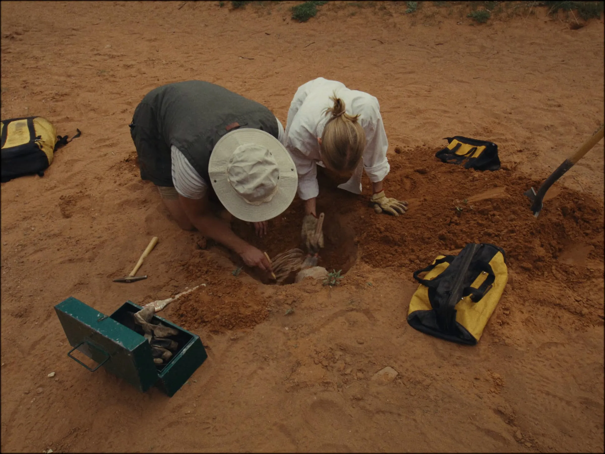 Two people kneel over a hole in red sand, uncovering a gauze-wrapped mummy, tools around them