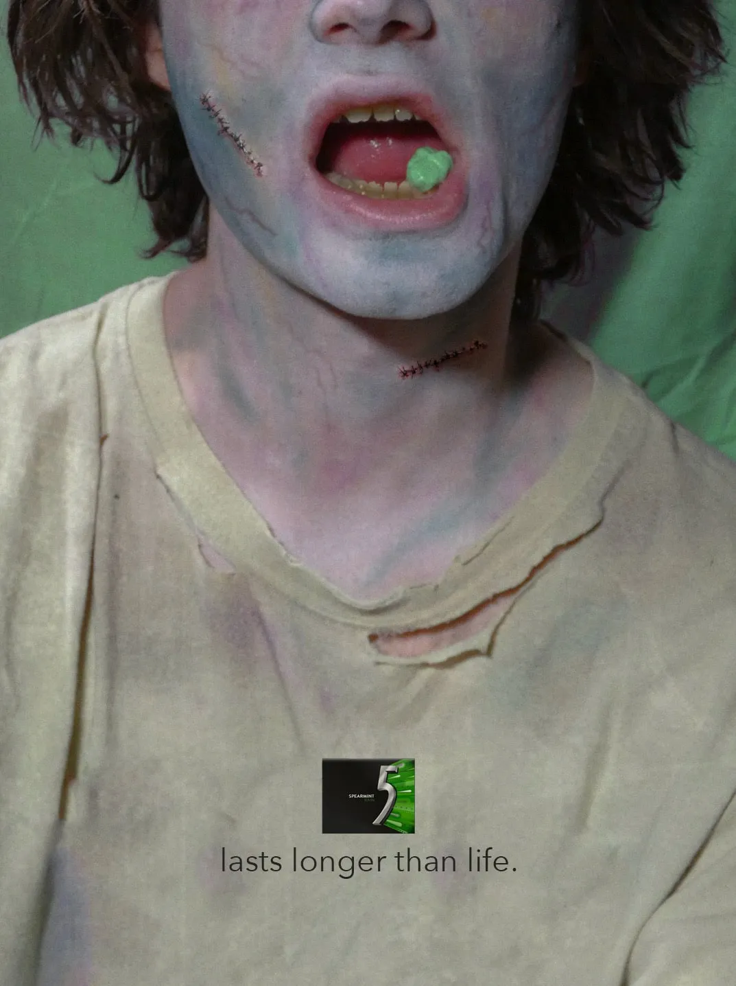 A zombie with stitched wounds on his cheek and neck shows a piece of green gum on his tongue. Under a pack of 5 Spearmint Rain: “lasts longer than life.”