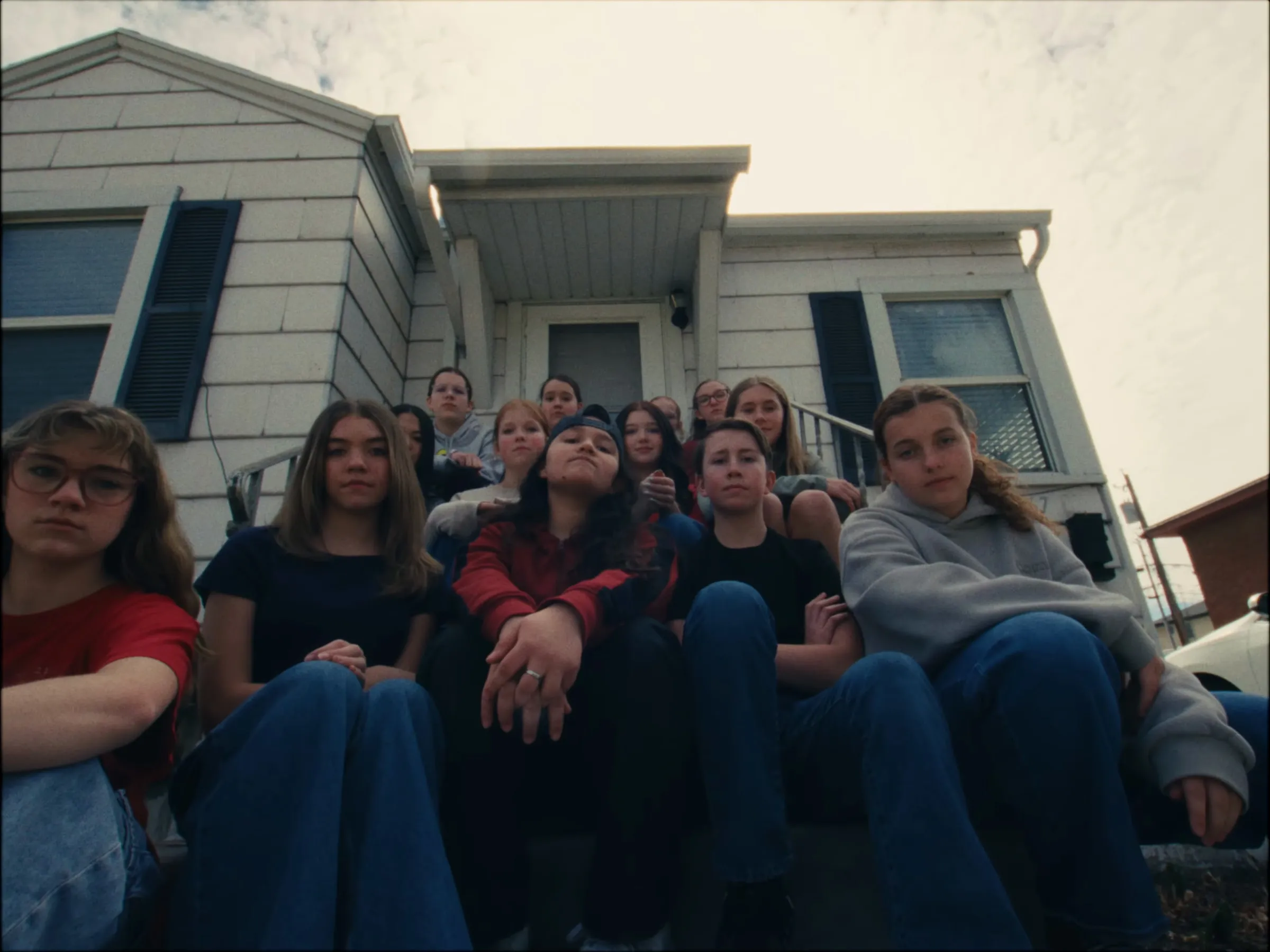 Shot from below, about a dozen teenagers packed together on the front steps of a white house, staring down into the lens