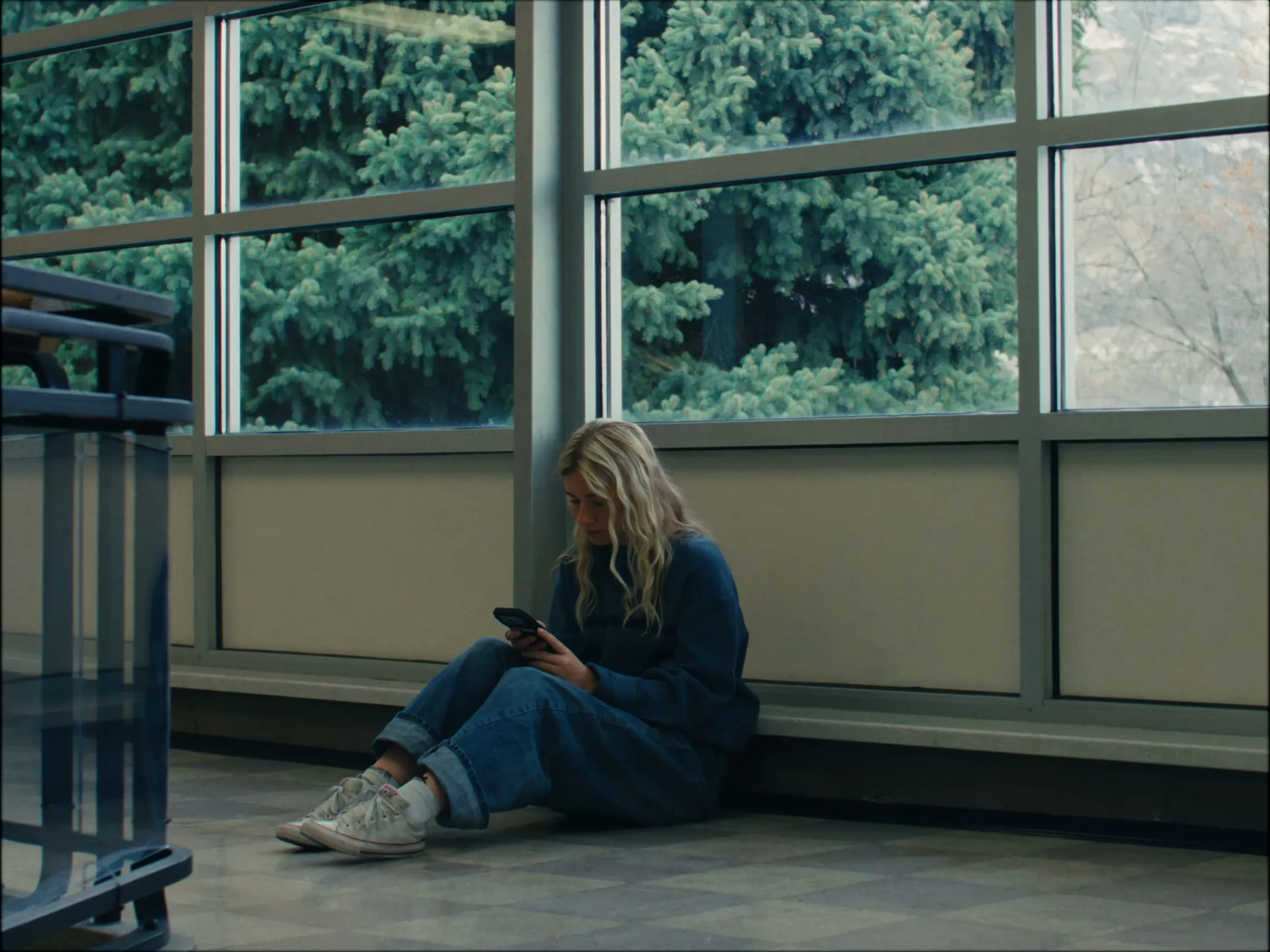A girl sits alone on the floor by a window, looking at her phone
