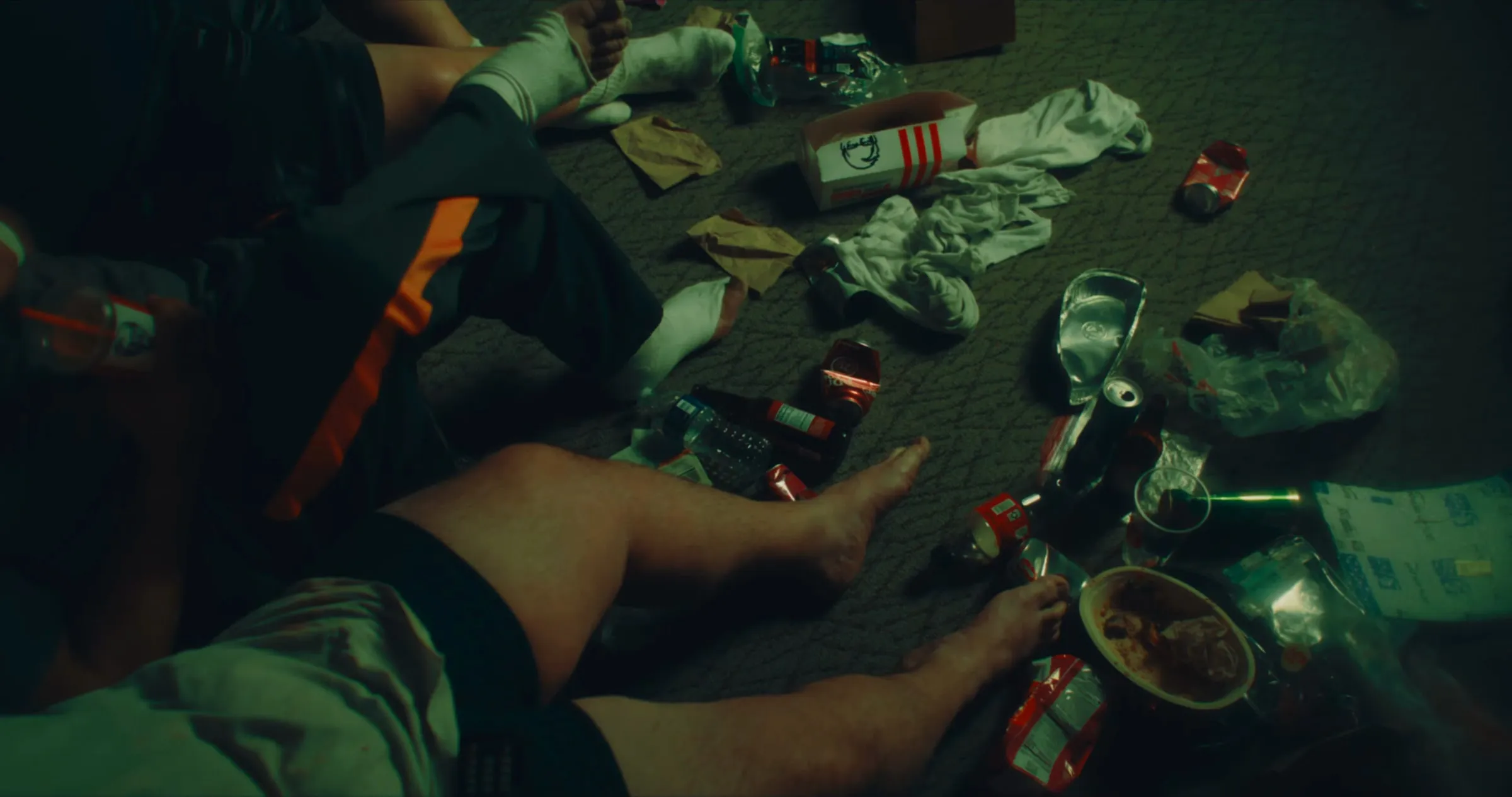 Legs and bare feet of several people on a carpet strewn with crushed cans, bottles and takeout trash