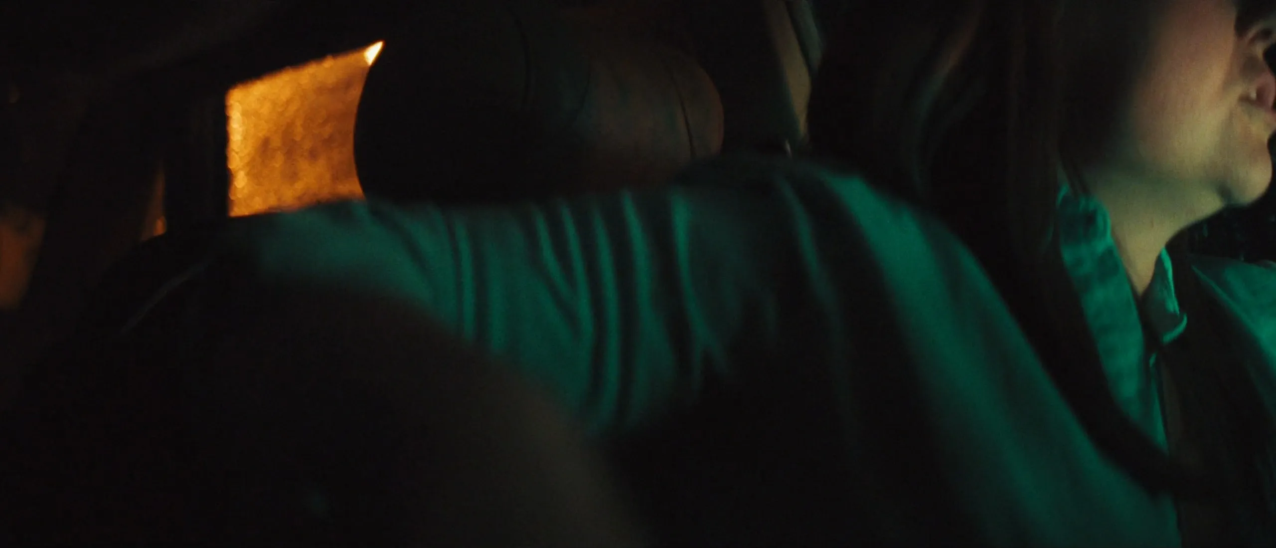 Inside a car at night, green light on a blurred arm in the foreground and on someone with long dark hair at the edge of the frame