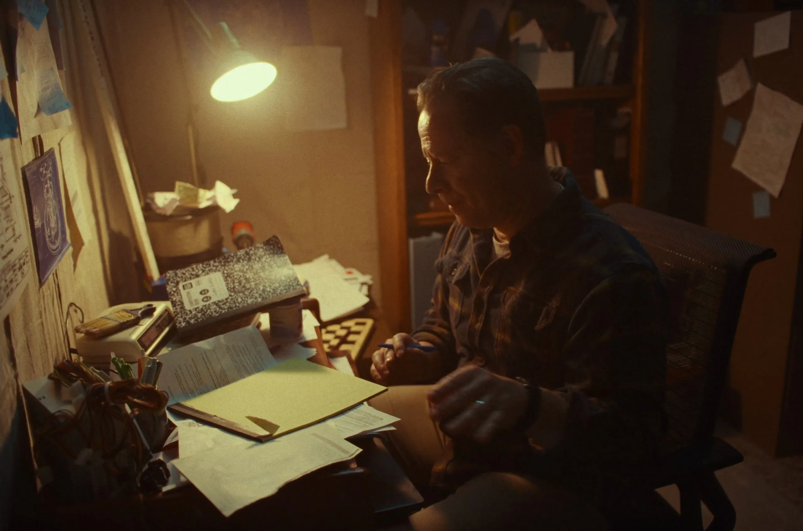A man works at a cluttered desk under a lamp