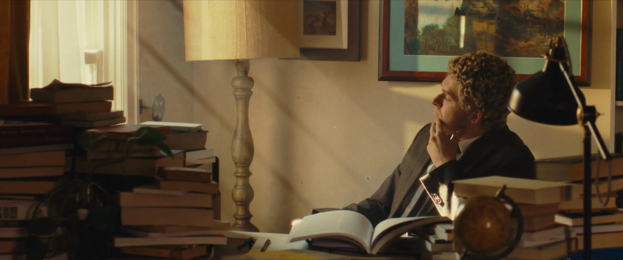 A man thinks at a desk piled with books