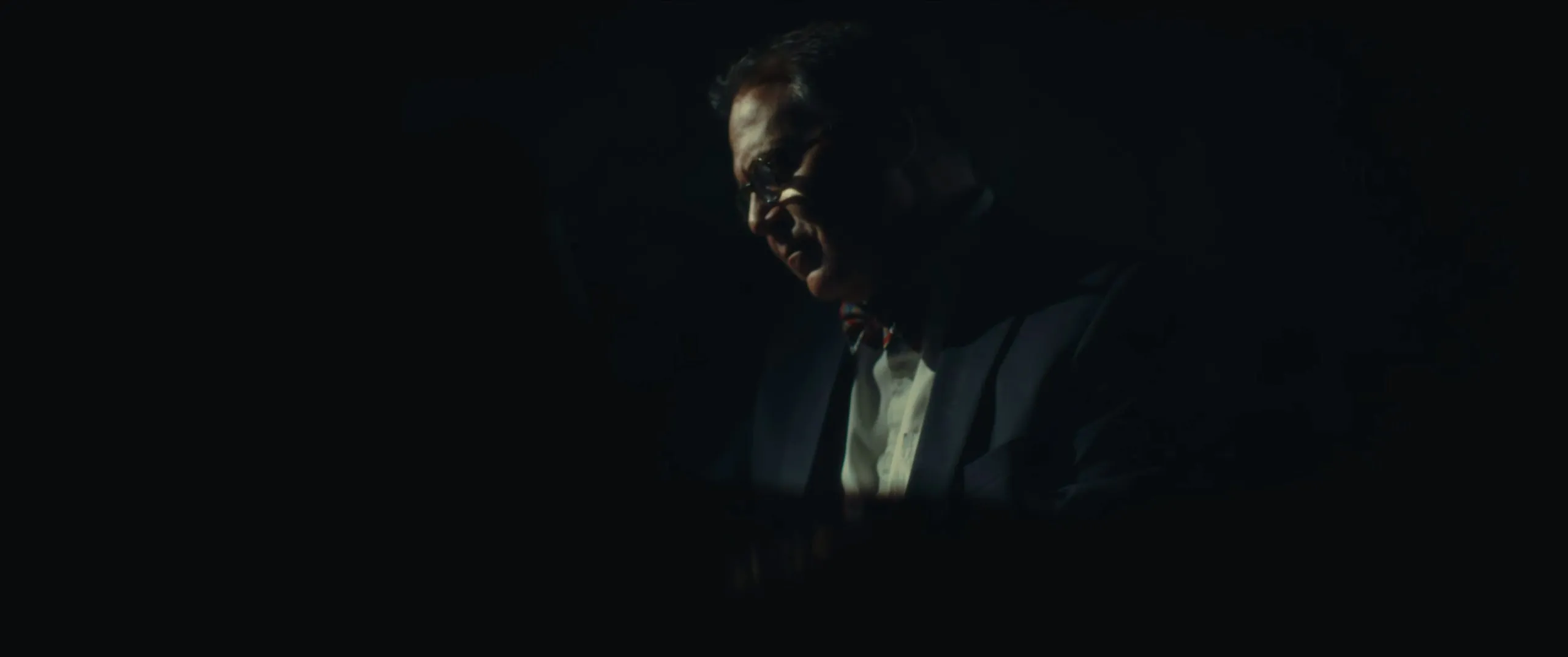 An older man in a suit sits in near darkness