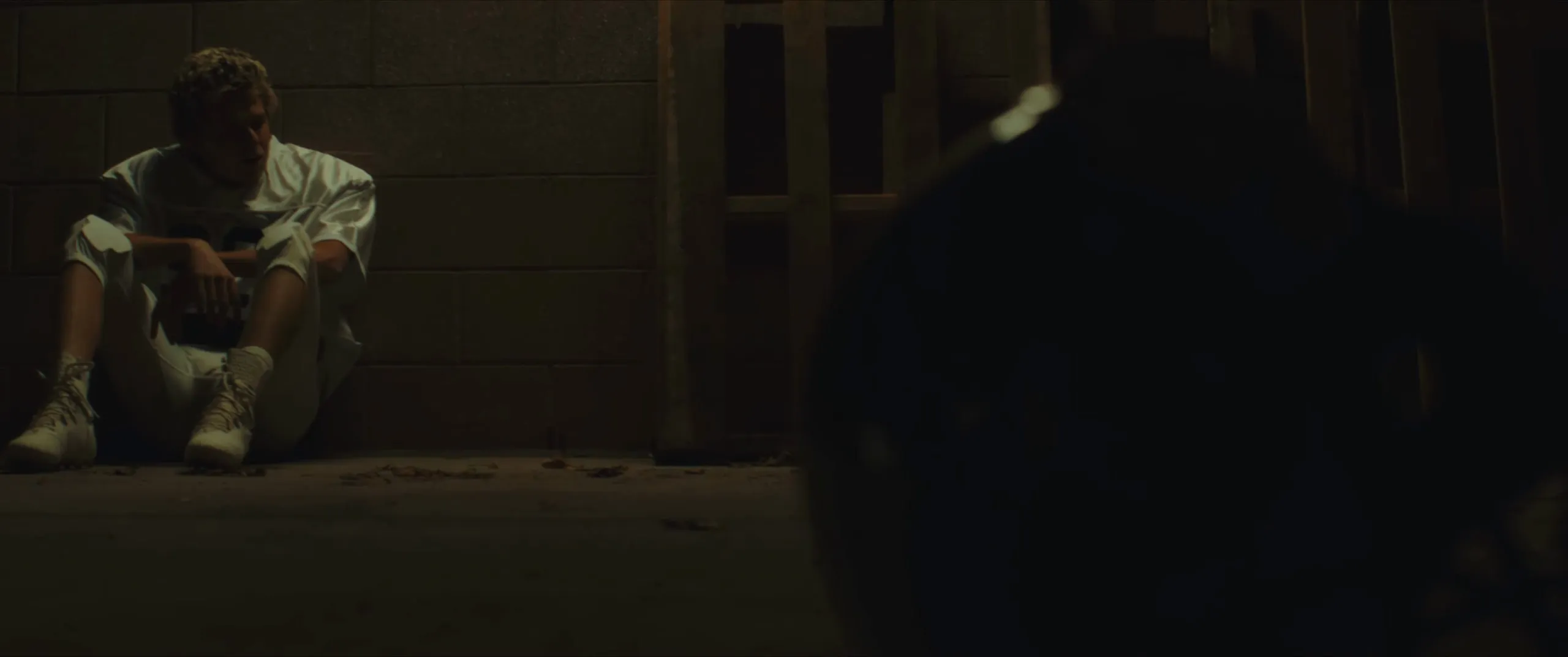 A young football player sits alone against a wall in the dark