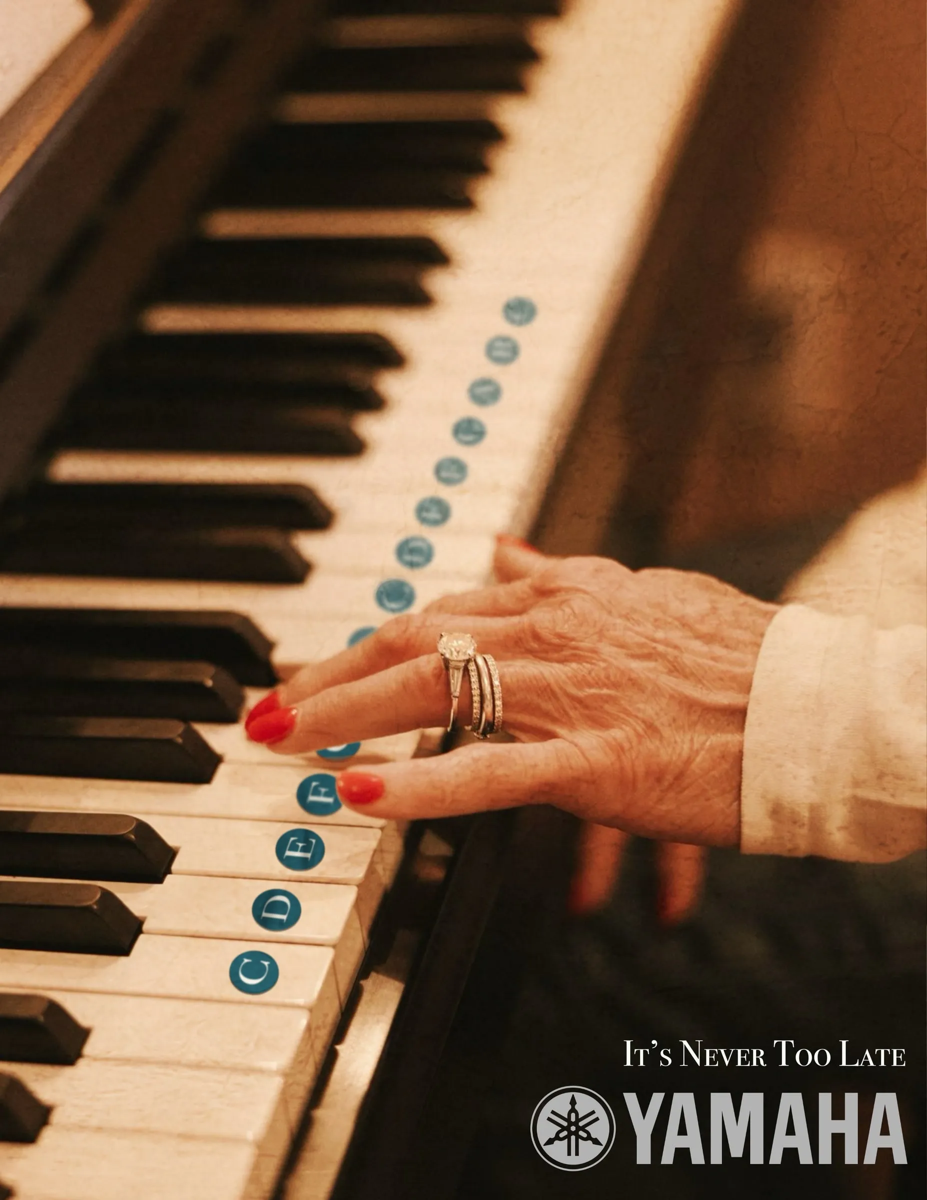 A wrinkled hand with red nails and a diamond ring rests on piano keys marked with note-name stickers. “It’s Never Too Late” with the Yamaha logo